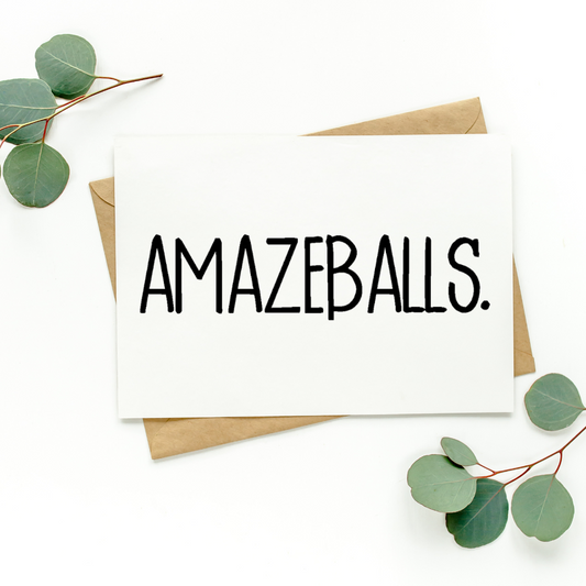 White greeting card with black text on top of kraft-colored envelope. Card reads, "Amazeballs.” Background of photo is white with green leaves in the top left and bottom right corners.