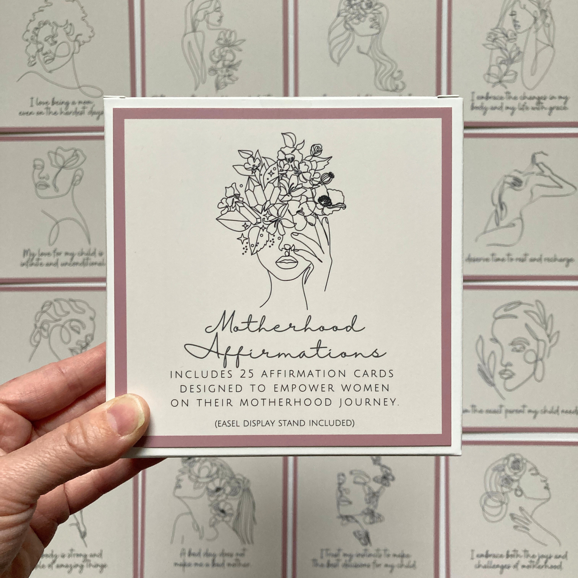 Hand holding a square affirmation card with line art illustration of a woman's face with flowers and the title 'Motherhood Affirmation Cards'. Other cards with similar design are visible in the background.