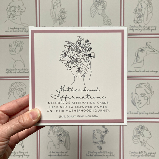 Hand holding a square affirmation card with line art illustration of a woman's face with flowers and the title 'Motherhood Affirmation Cards'. Other cards with similar design are visible in the background.