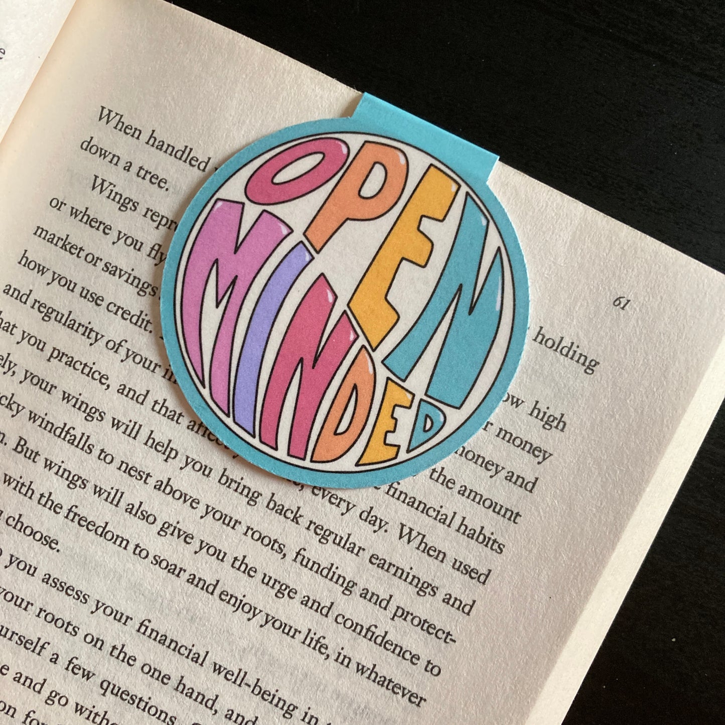 Round magnetic bookmark clipped around the page of a book. Bookmark features a teal background and the words Open Minded in bright colors
