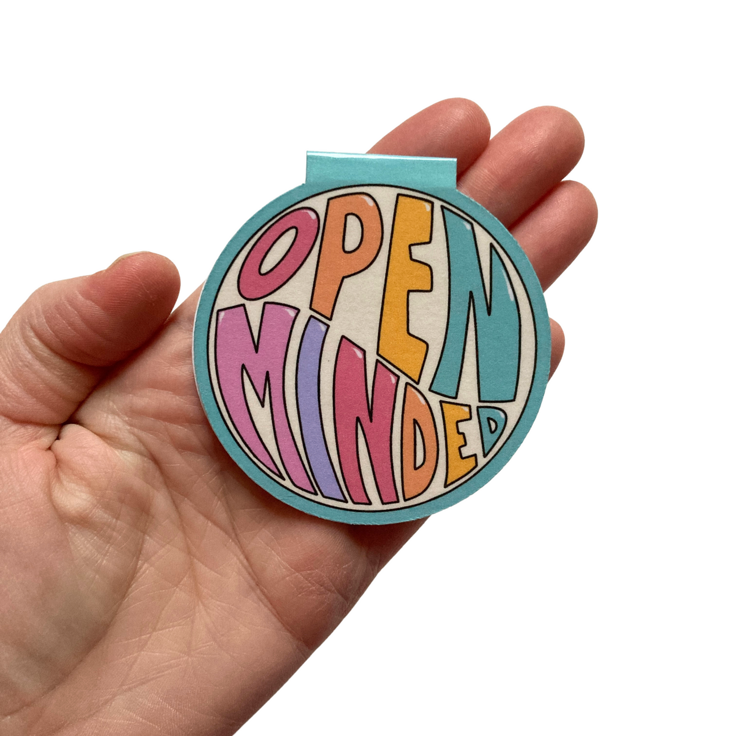 Round magnetic bookmark in palm of hand. Bookmark features teal background and the words Open Minded in large, colorful writing