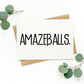 White greeting card with black text on top of kraft-colored envelope. Card reads, "Amazeballs.” Background of photo is white with green leaves in the top left and bottom right corners.