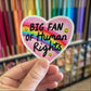 Heart-shaped sticker with 'BIG FAN OF Human Rights' text held in front of a colorful bookshelf.