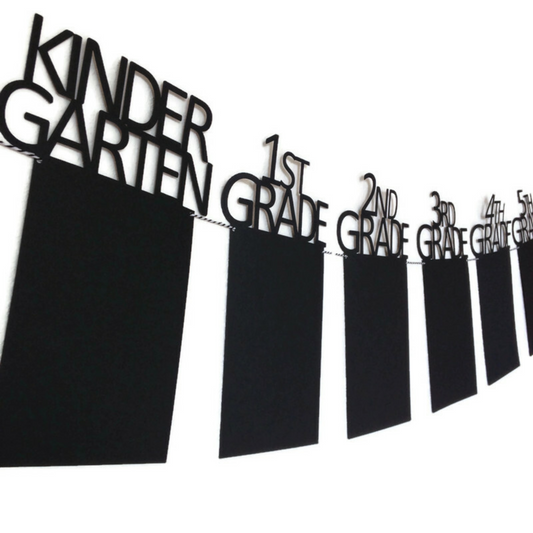 Black graduation banner with grades featuring kindergarten, first grade, and beyond. Strung on black and white twine and hanging on a white wall.
