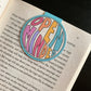 Round magnetic bookmark clipped around the page of a book. Bookmark features a teal background and the words Open Minded in bright colors