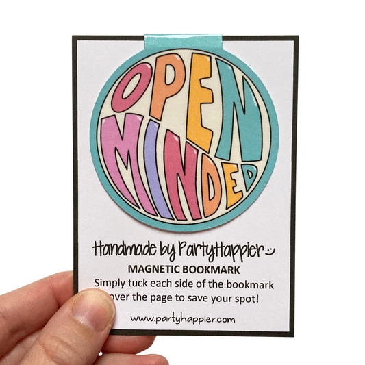 A hand is holding a round magnetic bookmark that is clipped onto a packaging card. Bookmark features a teal blue background with large, colorful letters that say Open Minded