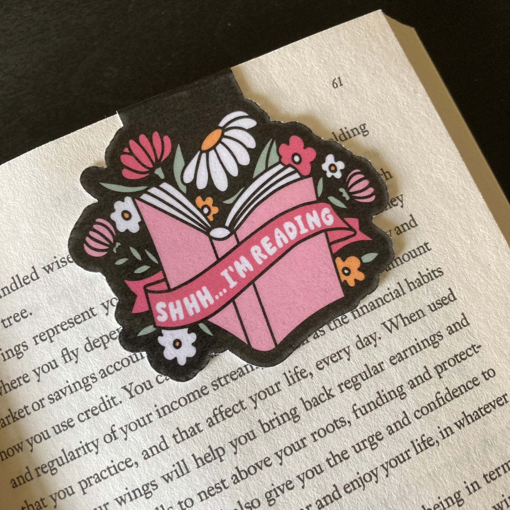 Magnetic bookmark clipped around the page of a book. Bookmark features a black background and a floral design with the words Shhh Im Reading across a graphic of a book