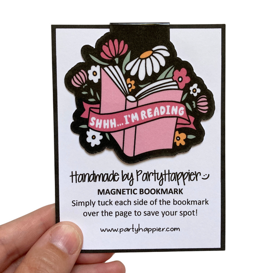 Hand holding magnetic bookmark that is clipped onto packaging card. Bookmark features a black background and floral design. Text on bookmark says Shhh Im Reading