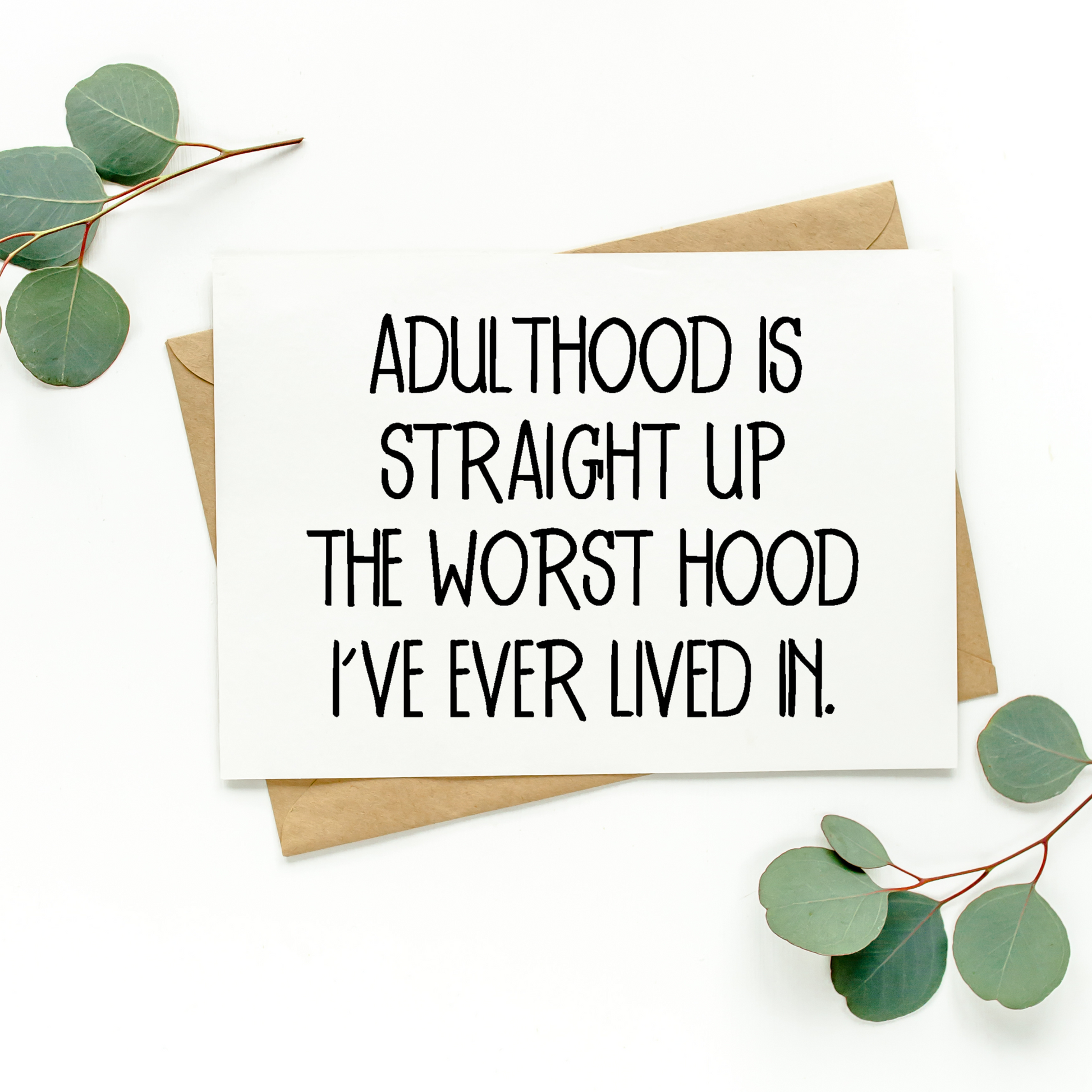White greeting card with black text on top of Kraft-colored envelope. Card reads "Adulthood Is Straight Up The Worst Hood I've Ever Lived In." Background of photo is white with green leaves in the top left and bottom right corners.