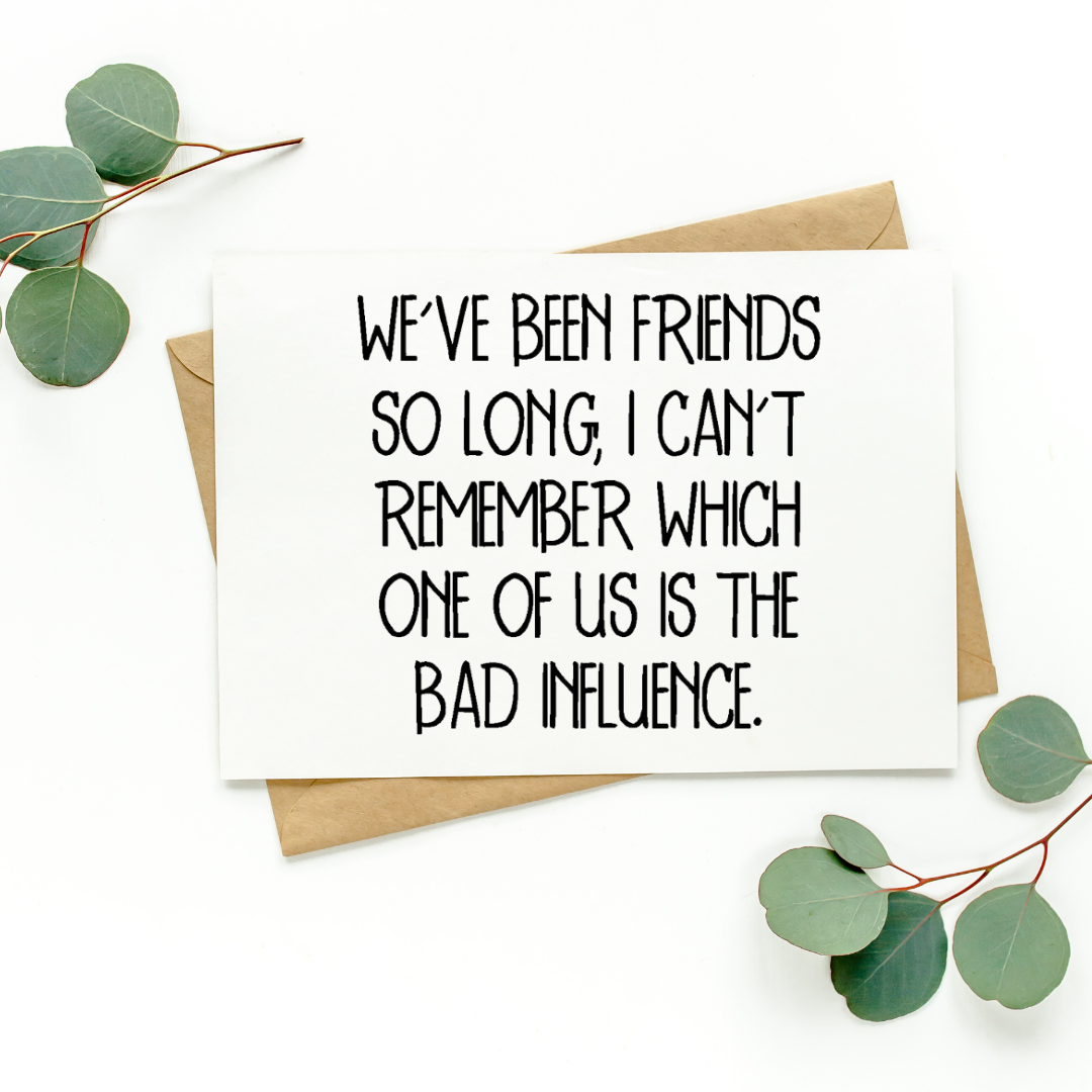White greeting card with black text on top of kraft-colored envelope. Card reads, "We’ve Been Friends So Long, I Can’t Remember Which One of Us Is The Bad Influence.” Background of photo is white with green leaves in the top left and bottom right corners.