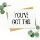 White greeting card with black text on top of kraft-colored envelope. Card reads, "You’ve Got This.” Background of photo is white with green leaves in the top left and bottom right corners.