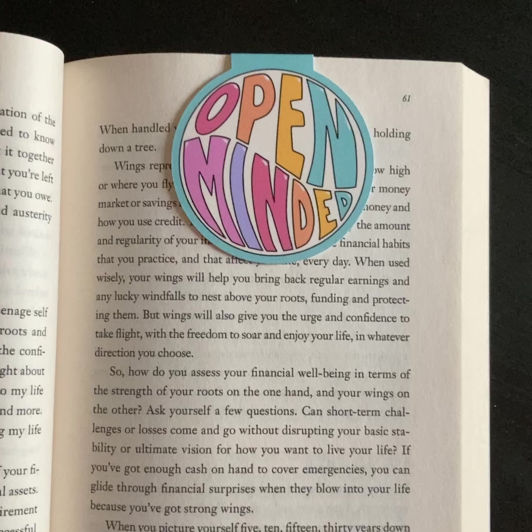 Video showing round magnetic bookmark clipped around the page of a book. The bookmark has a teal border and features the words Open Minded in large, colorful letters. The page turns and shows the same mirrored design on both sides.